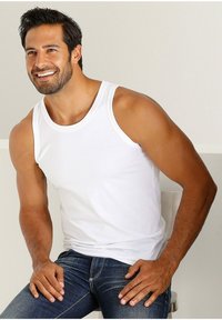 White cotton tank top with a round neckline and wide armholes, worn with dark blue jeans. The fabric is smooth and fitted.
