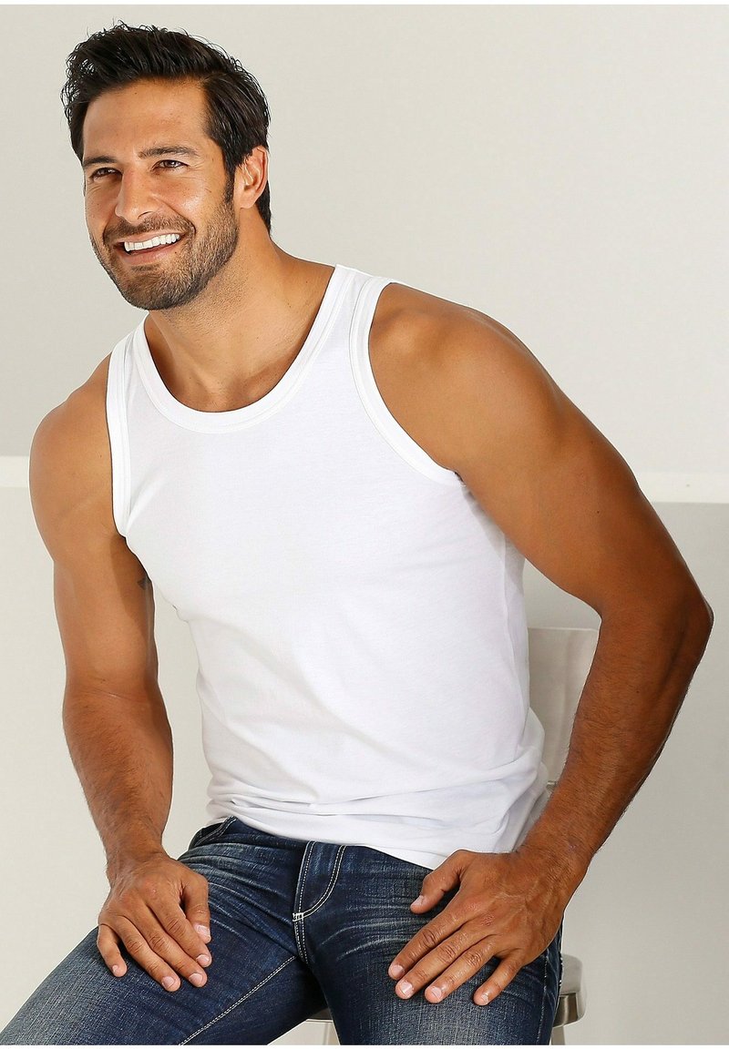 White cotton tank top with a round neckline and wide armholes, worn with dark blue jeans. The fabric is smooth and fitted.