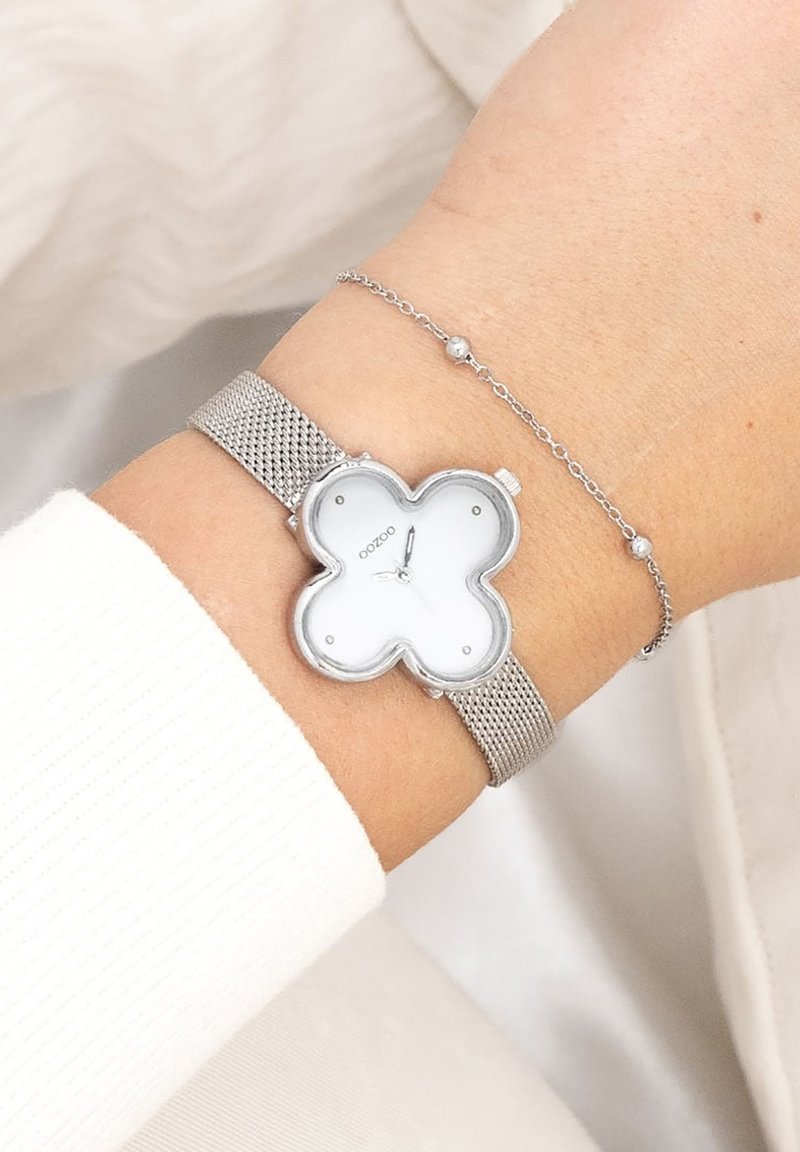 Montre à bracelet en maille argentée avec un cadran blanc en forme de fleur. Présente un design épuré, une texture lisse et des marqueurs d'heure minimalistes.