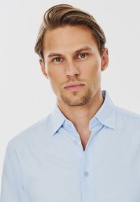 Jasnoniebieska koszula zapinana na guziki z kołnierzykiem, o subtelnej fakturze i płaskich guzikach, prezentowana na męskim modelu na jednolitym tle.