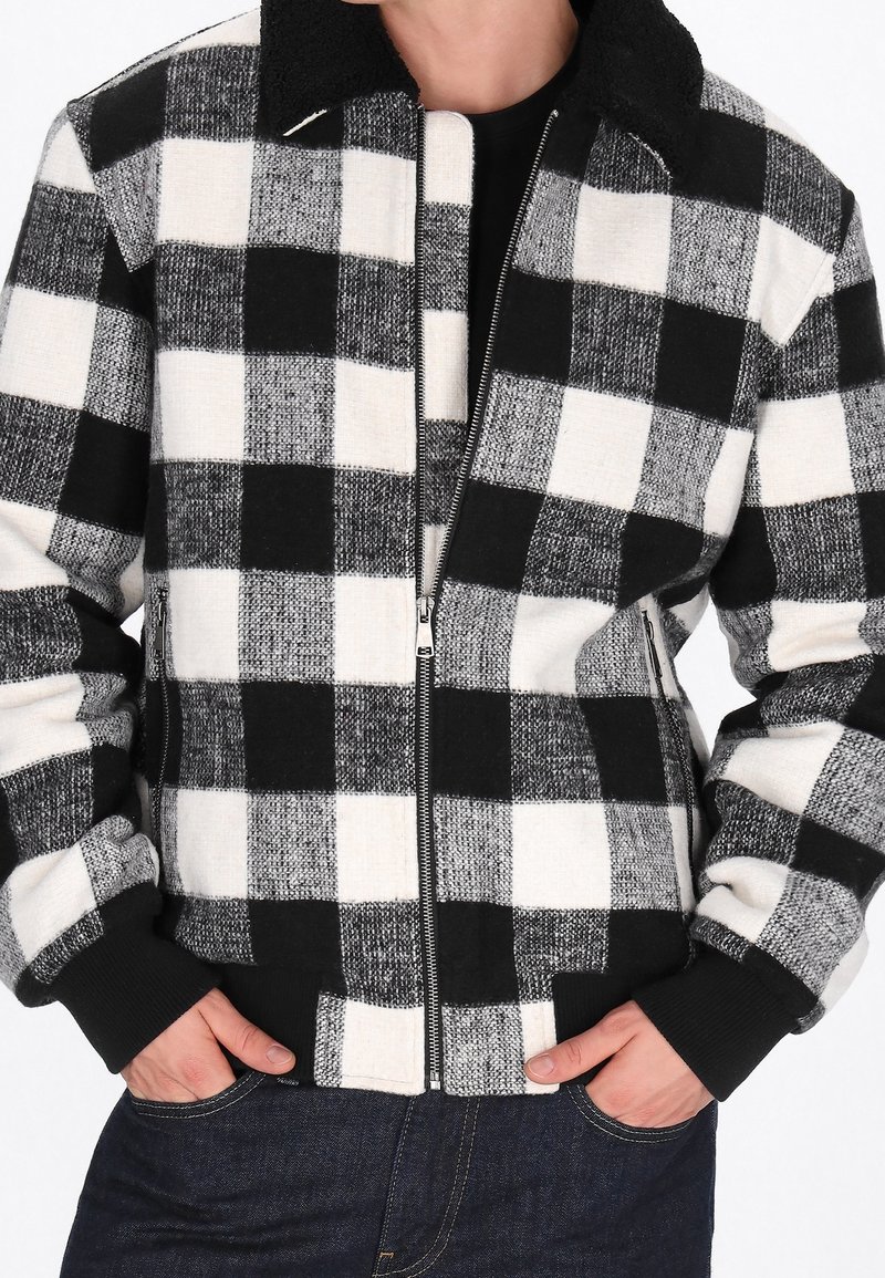 Veste zippée à carreaux noir et blanc avec poignets côtelés portée sur un t-shirt noir, mains dans les poches d'un jean bleu foncé.