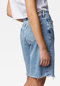 Donna che indossa pantaloni shorts in denim azzurro chiaro con vita alta e orli sfrangiati, insieme a una t-shirt bianca corta, in piedi contro uno sfondo bianco.