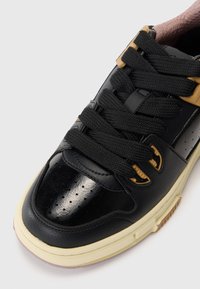 Black leather sneaker with a glossy finish, textured black laces, and yellow hardware accents. Features a cream-colored rubber sole.