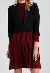 Chaqueta negra corta con solapas de satén sobre un vestido burdeos plisado. La tela es suave, con un diseño ajustado y mangas 3/4.
