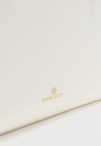Surface texturée beige clair avec petit logo et emblème dorés Anna Field centrés près du bord inférieur.
