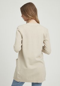 Beige kardigan s dlouhým rukávem vyrobený z měkkého pleteného materiálu, má kulatý výstřih a boční kapsy, s volným střihem a žebrovanými manžetami.