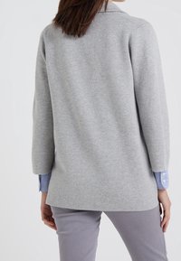 Grå stickad tröja med klassisk krage, med 3/4-långa ärmar och en avslappnad passform. Under syns ärmkuvor av en blå och vit randig skjorta.