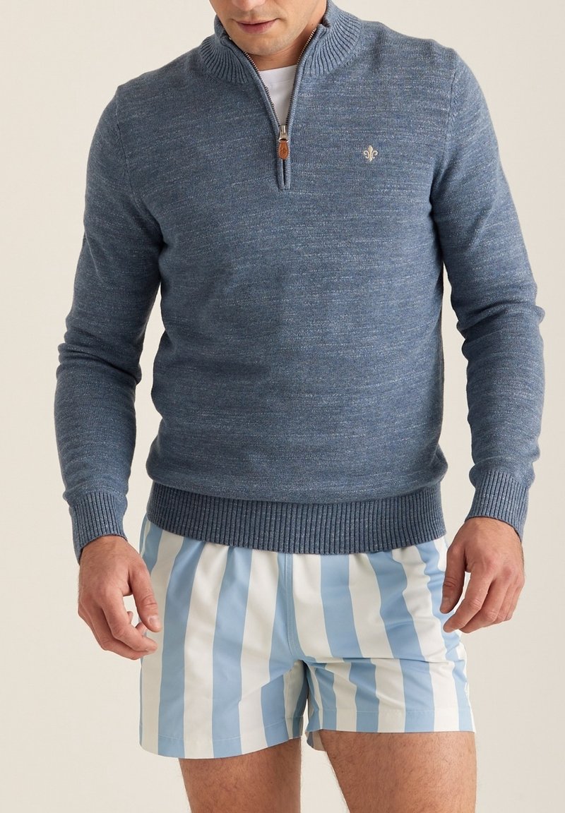 Homme portant un pull bleu à fermeture éclair quart de zip avec des poignets côtelés et un short rayé vertical bleu et blanc, debout sur un fond neutre.