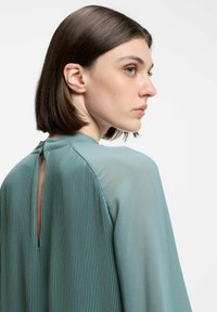 Teal pleated blouse featuring a keyhole back detail, long sleeves, and a smooth, textured fabric.