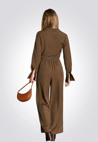 Bruine, lichte jumpsuit met een kraag, belmouwen en een elastieke taille. Gecombineerd met een bruine, gebogen handtas en enkellaarsjes.