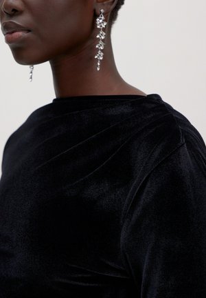 Nahaufnahme einer Person, die ein schwarzes Samtoberteil und lange silberne, hängende Ohrringe mit kristallähnlichen Details trägt.