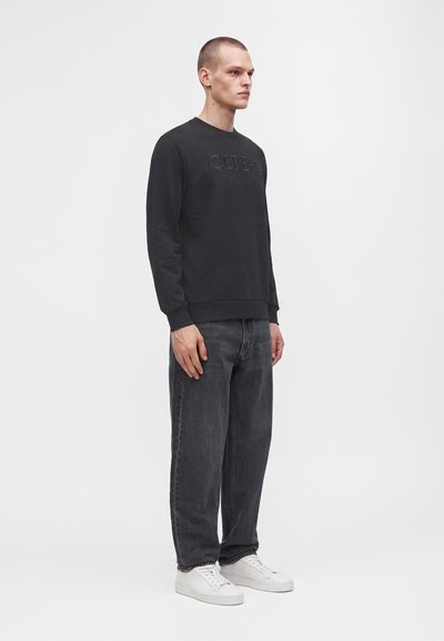 Homme debout de profil portant un sweat-shirt noir, un jean gris foncé et des baskets blanches sur un fond gris uni.