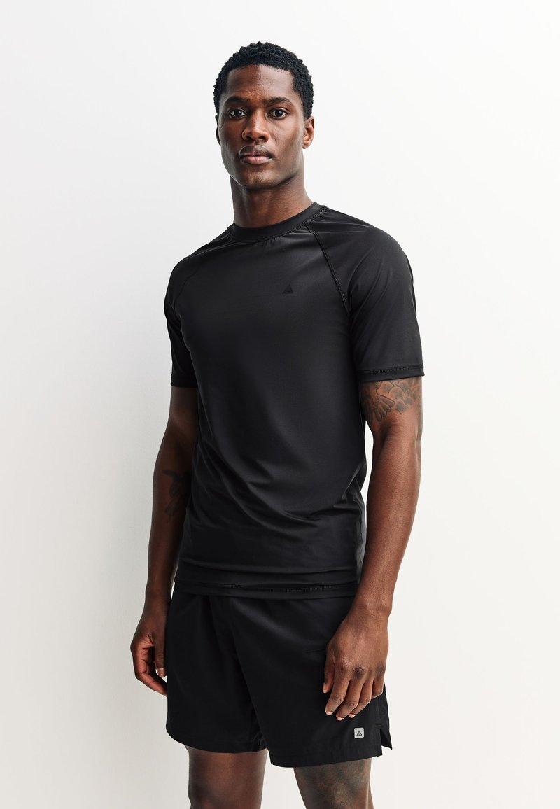Modèle masculin portant un haut de sport à manches courtes noir et un short assorti, debout devant un fond clair uni, faisant face à l'avant.