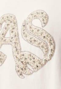 Broderie perlée complexe avec des perles blanches et de petites pierres précieuses argentées formant les lettres "A" et "S" sur un tissu crème.