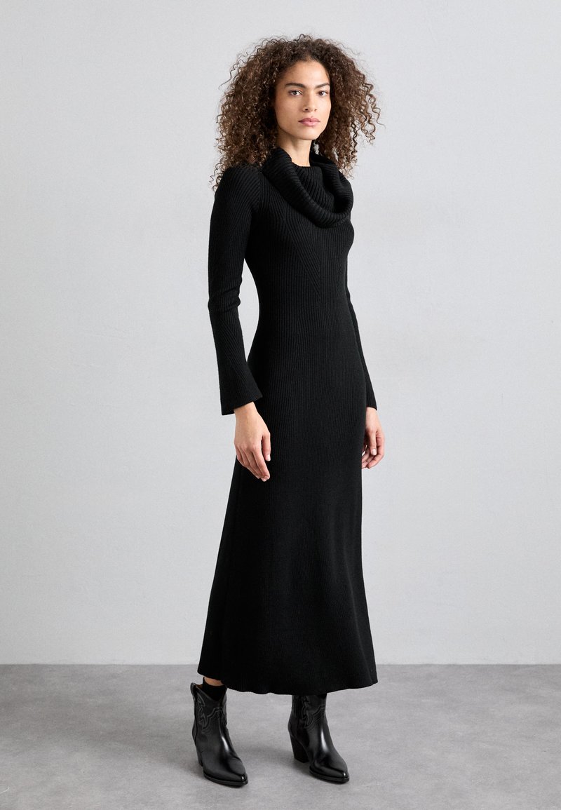 Black ribbed knit dress with a cowl neck, long sleeves, and a flowing skirt. Paired with black ankle boots featuring a decorative pattern.