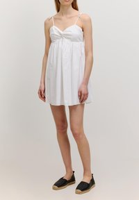 Witte mouwloze jurk met een gedraaid bovenstuk, gestructureerde stof en een vloeiende rok. Gecombineerd met zwarte espadrille-achtige schoenen.