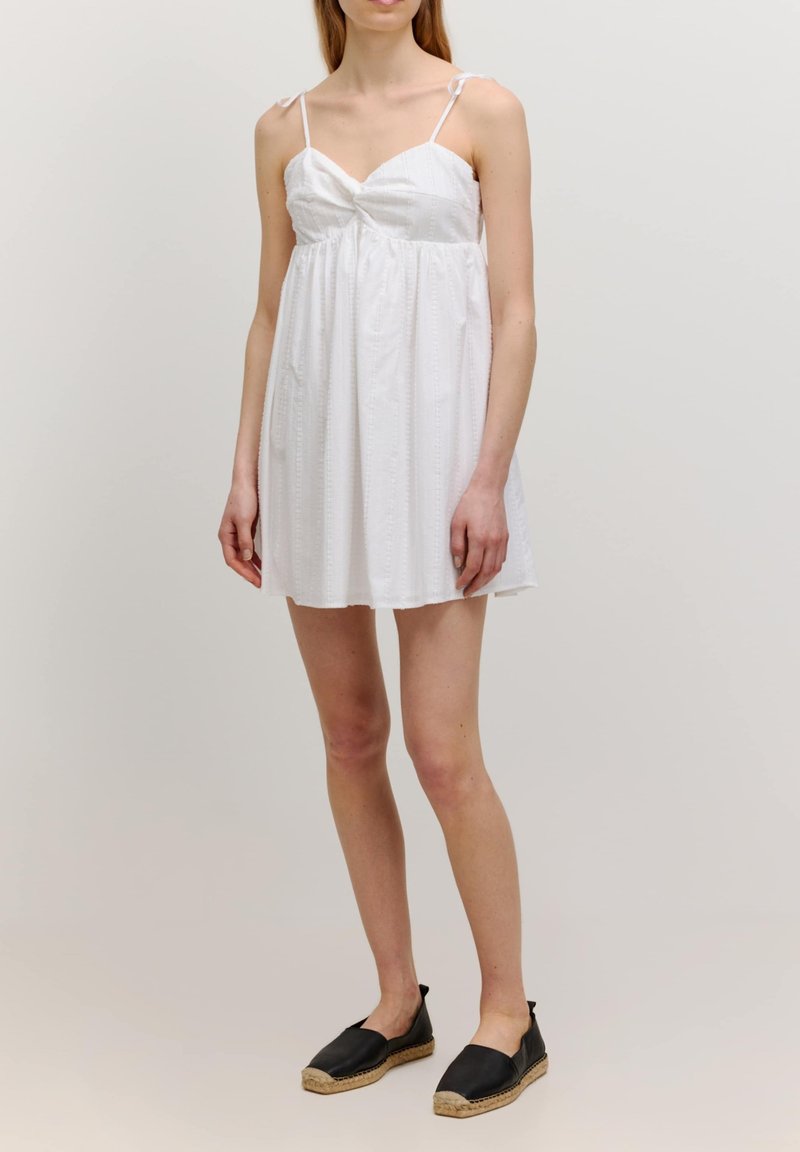 Witte mouwloze jurk met een gedraaid bovenstuk, gestructureerde stof en een vloeiende rok. Gecombineerd met zwarte espadrille-achtige schoenen.