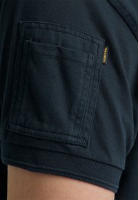 Dunkelblaues Poloshirt mit strukturierter Oberfläche, ausgestattet mit einer aufgesetzten Tasche auf dem Ärmel und einem gelben Markenlabel.