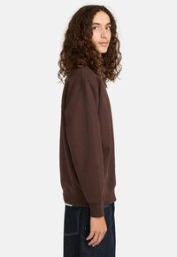 Felpa marrone realizzata in morbido cotone, dotata di maniche lunghe, polsini a costine e una vestibilità rilassata. Dettagli verdi notevoli appaiono sul retro.