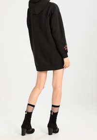 Henkilö, jolla on päällä musta ylisuuri huppari, jossa on punainen tähtiommel hihassa, kalastajaverkkosukat ja mustat korkeat nilkkakengät, seisoo sivuttain.