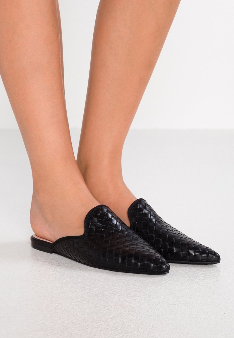 Pieds portant des chaussures mule tissées noires à bout pointu, sans bride, sur un sol clair avec un fond blanc.