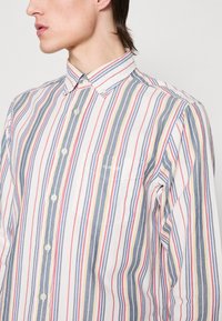 Camisa de botones a rayas con rayas verticales en azul, rojo y amarillo sobre un fondo blanco, con un bolsillo en el pecho y cuello camisero.