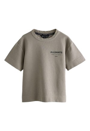 Junge trägt ein übergroßes olivgrünes AllSaints-T-Shirt, schwarze Jeans und weiße Sneaker und steht mit einer Hand in der Tasche.