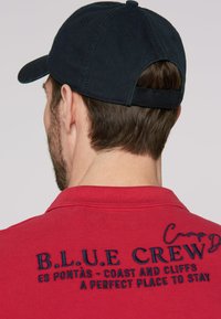 Red polo shirt with navy embroidery reading “B.L.U.E CREW” and text below. Features a navy baseball cap. Cotton material, short sleeves.