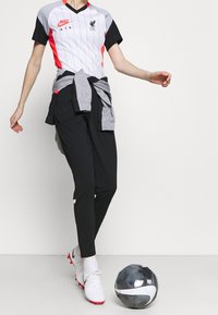 Koszulka Nike Liverpool FC w szarym kolorze z czerwonymi akcentami, zestawiona z czarnymi spodniami sportowymi. Obok na ziemi leży piłka nożna.