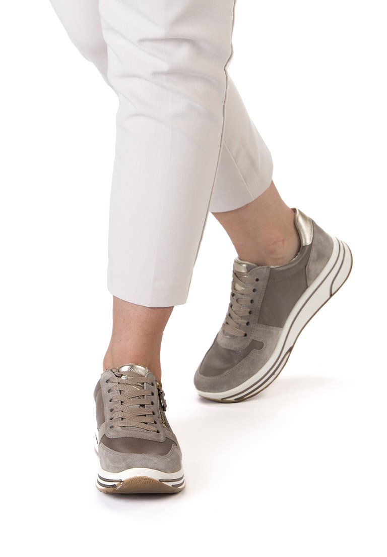 Person trägt beige Freizeitsneaker mit Plateausohlen und weiße Hose, steht und macht einen Schritt nach vorne vor weißem Hintergrund.