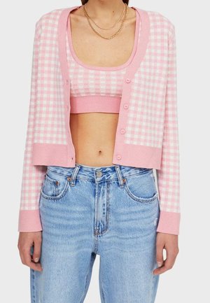 Vrouw draagt een roze-wit geruite cropped cardigan en een bijpassende crop top, gecombineerd met een lichtblauwe spijkerbroek met hoge taille en gouden kettingen.