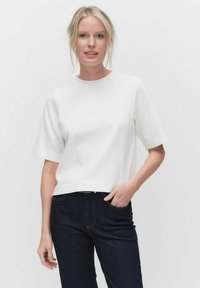 Valkoinen lyhyt toppi, jossa on pyöreä kaula-aukko ja lyhyet hihat, valmistettu sileästä kankaasta. Yhdistetty tummansinsisiin farkkuihin. Yksinkertainen malli, ilman kuvioita.