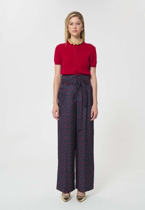 Femme portant un haut rouge à manches courtes boutonné, un pantalon large bleu marine avec un motif rouge et une ceinture à la taille, un collier en or, des boucles d'oreilles et des talons pointus dorés.