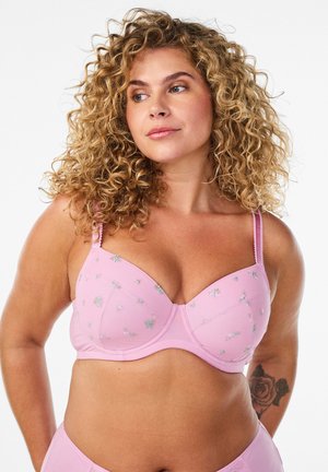 Kvindelig med krøllet hår og lys hud, iført en lyserød blomstret bh, kigger til siden, med en rose-tatovering på venstre arm.