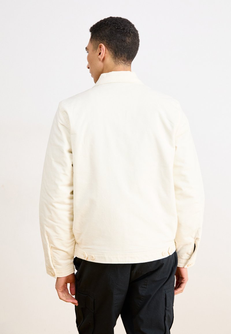 Veste de couleur crème avec une texture lisse, manches longues et boutons aux poignets, associée à un pantalon cargo foncé. Vue de dos.