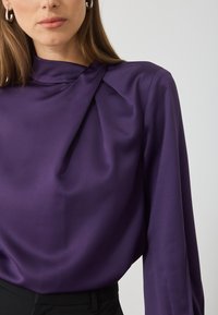 Bluză din satin mov cu decolteu plisat, mâneci lungi și o croială adaptată, asortată cu pantaloni negri cu talie înaltă. Textură netedă.