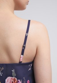 Námořnické květinové šaty s tenkými ramínky; květinový vzor obsahuje růžové a bílé květiny. Hladká tkanina s mírným leskem. Pohled zezadu.