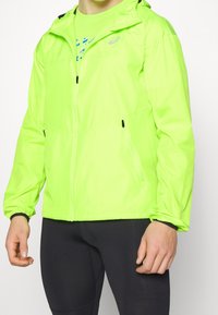 Veste légère vert néon avec capuche, poches zippées, poignets élastiques et tissu lisse, résistant à l'eau.