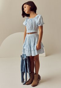 Világoskék virágos rövid felső puffos ujjakkal és hozzáillő, réteges szoknya. Barna bokacsizma bojtos részletekkel. Farmerruha a karon átdobva.