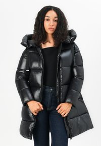 Zwarte pufferjas met een glanzende afwerking, een oversized kap en gestikte panelen. Voorzien van een ritssluiting aan de voorkant en zijzakken, gestyled met donkere spijkerbroek.