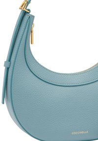 Jasnoniebieska skórzana torebka o zaokrąglonym kształcie, złote elementy ozdobne, detal na suwaku oraz fakturowane wykończenie. Oznaczona marką "Coccinelle".
