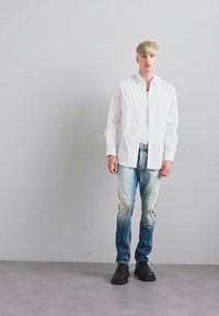 Jeune homme debout, portant une chemise blanche à boutons, un jean bleu clair délavé avec un patch, et des chaussures noires, devant un mur gris uni.