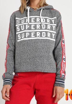 Osoba nosząca szarą bluzę z kapturem Superdry z białymi sznureczkami i czerwonymi paskami na rękawach, do czerwonych spodni.