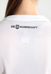 Witte sporttrui met een meshstructuur, waarin "DIE MANNSCHAFT" zwart is gedrukt op de bovenrug, vergezeld van een logo met sterren.