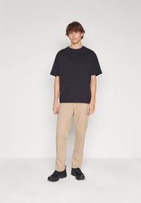 Jonge man die staat, gekleed in een zwart T-shirt met korte mouwen, beige cargobroek en zwarte sneakers, tegen een eenvoudige witte achtergrond.