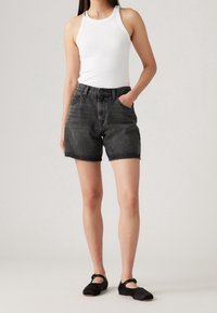 Pantalones cortos de mezclilla negra con cintura alta y dobladillo deshilachado, combinados con una camiseta sin mangas ajustada blanca. La modelo lleva unos zapatos de charol negros tipo Mary Jane.