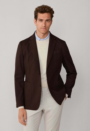 Mann trägt dunkelbraunes Sakko, cremefarbenen Pullover, hellblaues Hemd und hellgraue Hose, steht mit einer Hand in der Tasche.