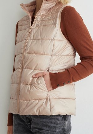 Femme portant un gilet matelassé beige sans manches par-dessus une chemise à manches longues couleur rouille, main dans la poche, debout devant un fond blanc.