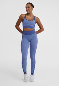 Reggiseno sportivo blu e leggings a vita alta in un tessuto leggero e testurizzato. Dotati di inserti in rete e design aderente. Sneakers bianche.