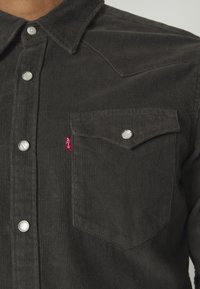 Gros plan d'une chemise en velours côtelé foncé avec des boutons-pression blancs et une poche poitrine à rabat avec une étiquette rouge Levi's.